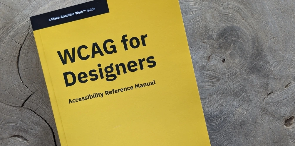 Paperback book with a bright yellow cover and black text with the title, 'WCAG for Designers: Accessibility Reference Manual,' series name, 'A Make Adaptive Work Guide,' and author name 'Stacey Swinehart Ganderson.' The book is on a pine-colored, smooth wood stump tabletop.