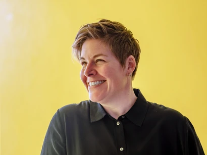 Woman with short hair, wearing a black collared and button-down shirt, looking to the left and smiling against a bright yellow background.