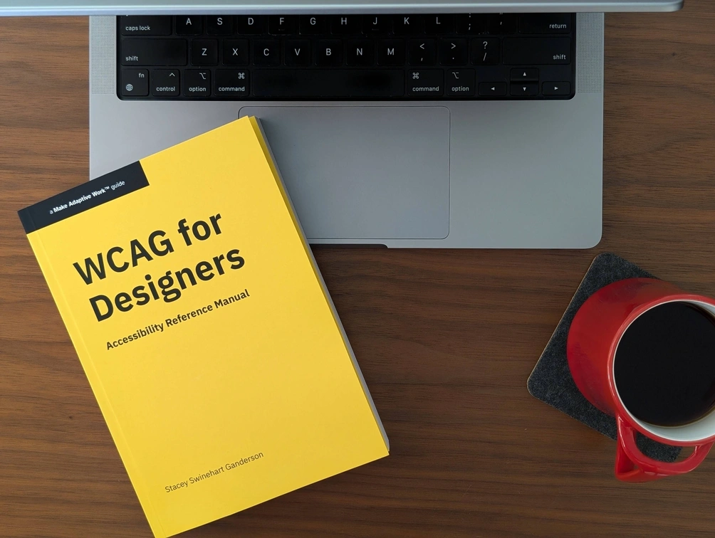 Paperback book with a bright yellow cover and black text with the title, 'WCAG for Designers: Accessibility Reference Manual,' series name, 'A Make Adaptive Work Guide,' and author name 'Stacey Swinehart Ganderson.' The book is on a partially open laptop next to a red mug with tea on a walnut-colored tabletop.